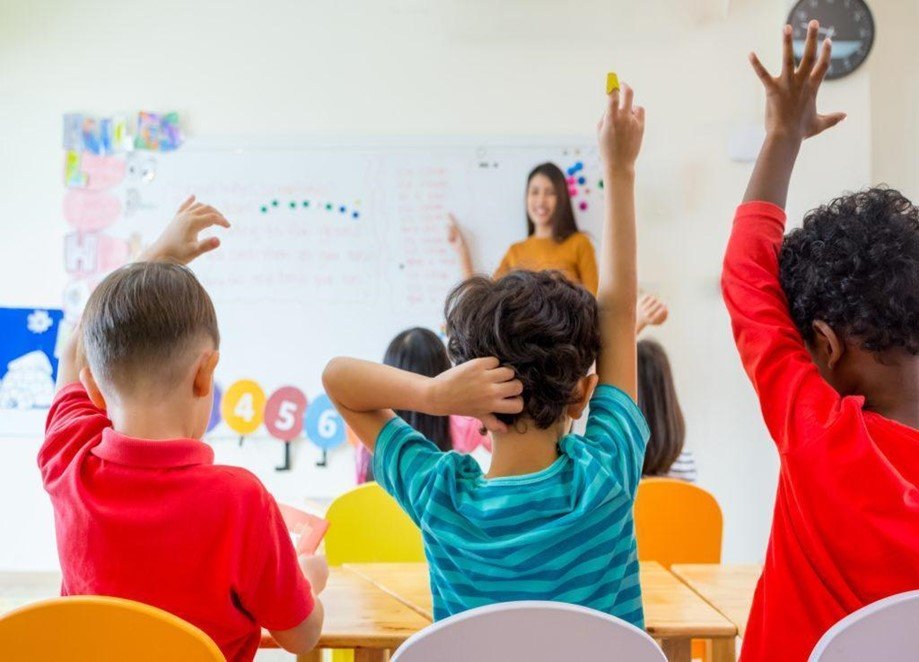 Elementary kids with hands raised, facing teacher at whiteboard